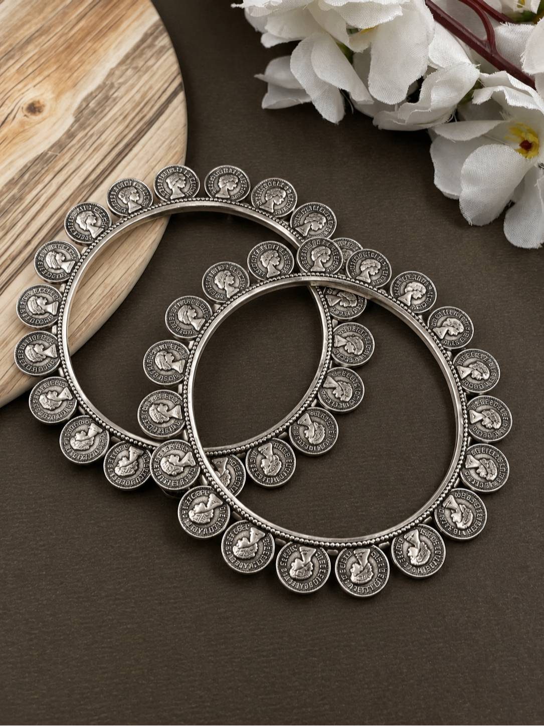 Pair of oxidised silver coin kadas laid flat on a dark surface beside white flowers and a wooden prop