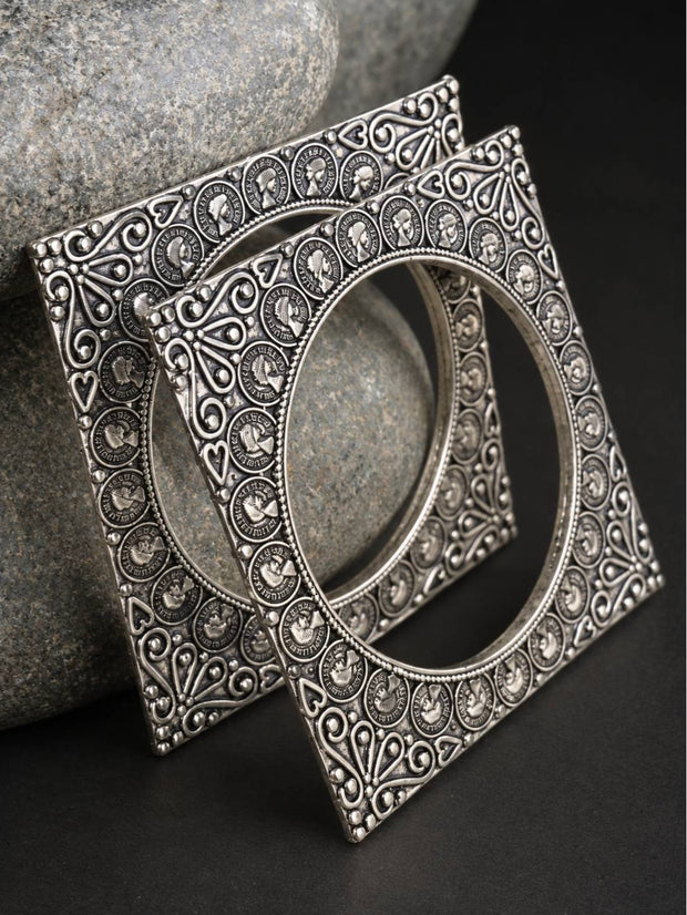 Two oxidised silver square kadas with coin motifs leaning against grey stones on a dark background