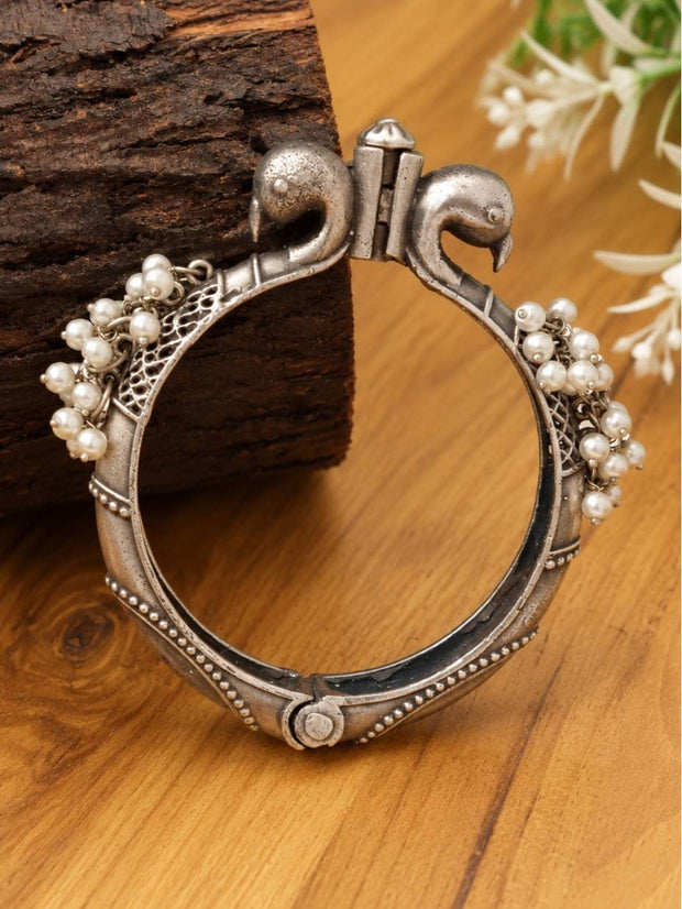 Oxidised silver peacock kada with pearl bead clusters resting against a dark wood block, side view showing dual bird-head clasp