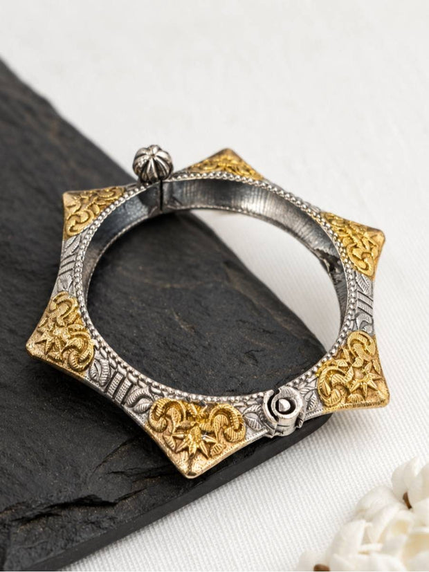 Heptagonal dual-tone oxidised silver floral kada resting on a dark slate surface with white flowers in background