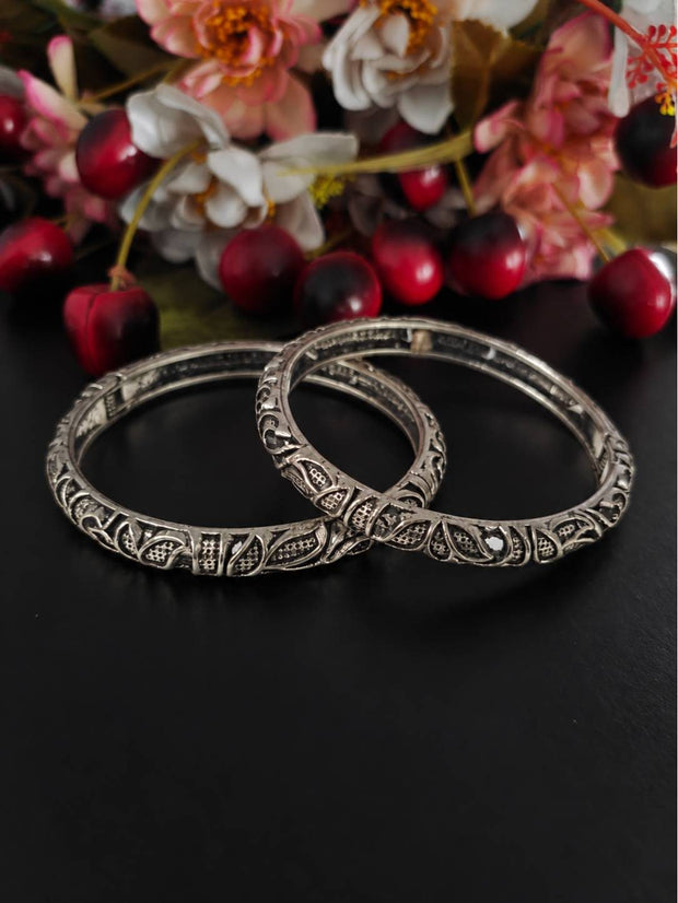 Two oxidised silver paisley bangles laid flat against a dark surface with red berries and flowers in background