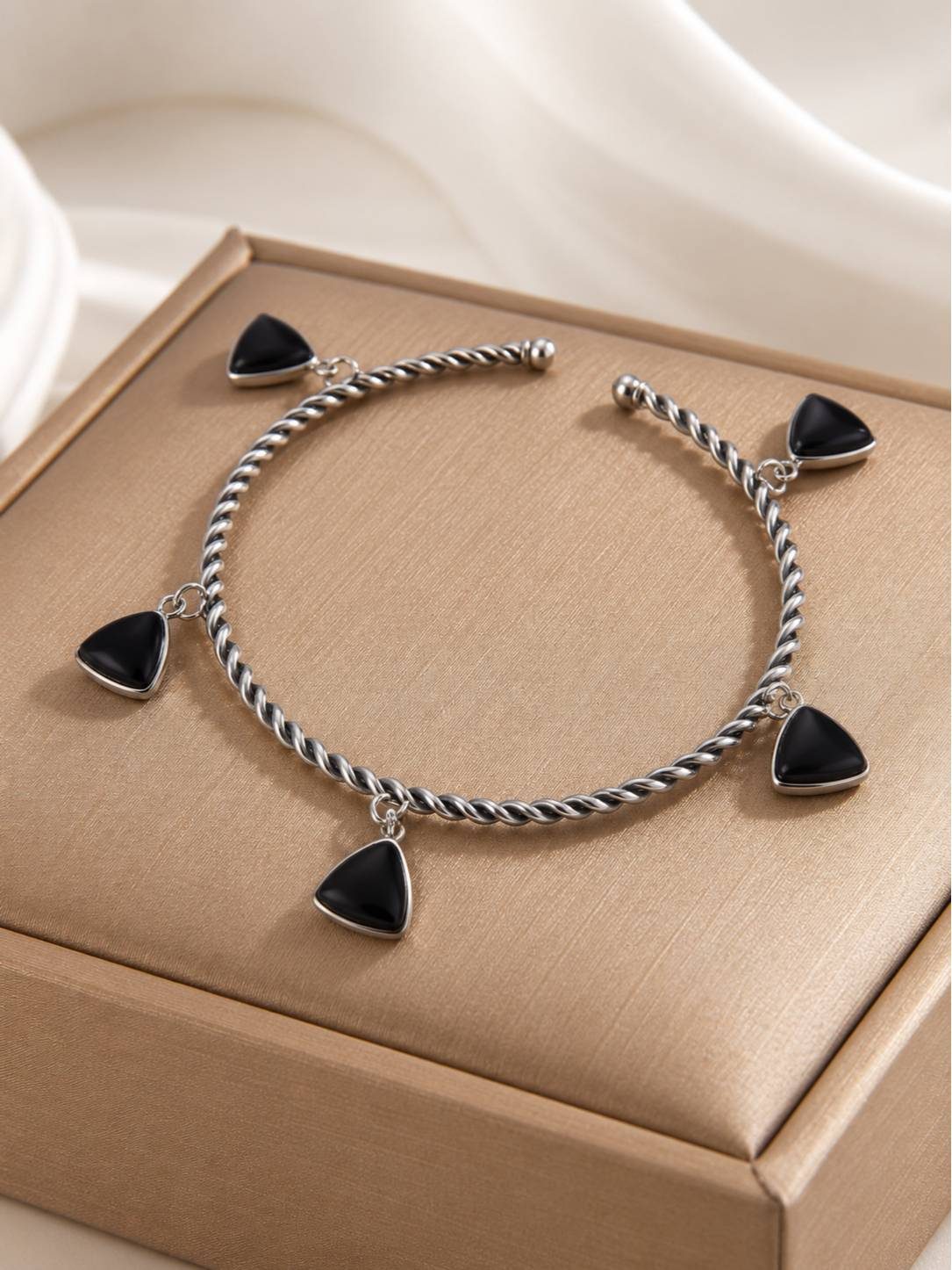 Oxidised silver twisted cuff bracelet with five black triangle charm drops laid flat inside a gold jewellery box