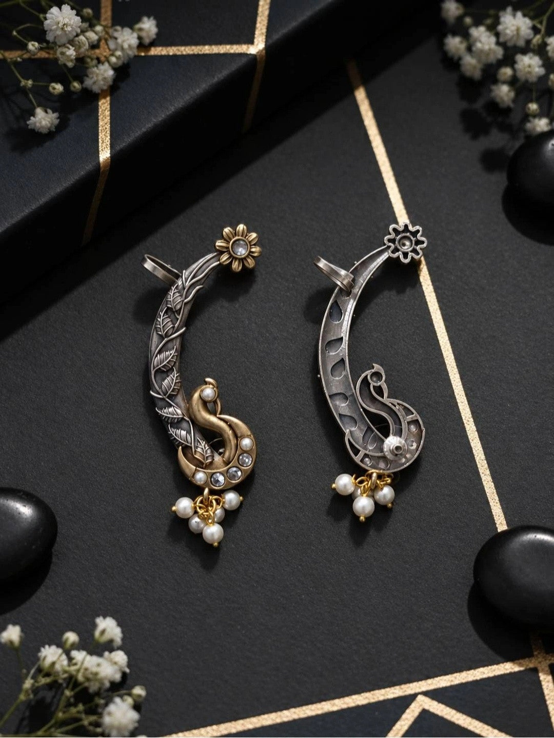Pair of oxidised dual-tone peacock ear cuffs with pearl drops laid flat on black surface
