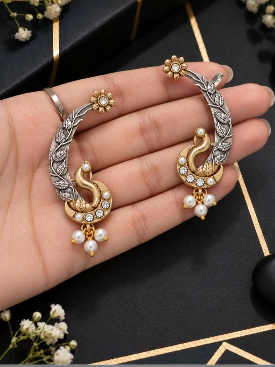 Both peacock ear cuffs held in an open palm showing gold-tone peacock and silver leaf engraving