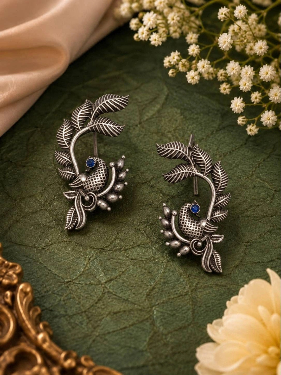 Pair of oxidised silver peacock ear cuff earrings laid flat on dark green leather surface