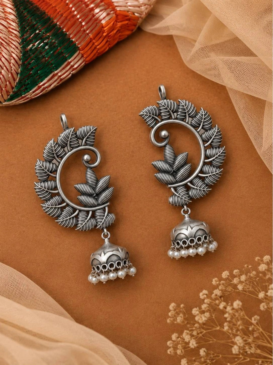 Two oxidised silver leaf jhumka earrings laid flat on a warm terracotta surface with dried flowers