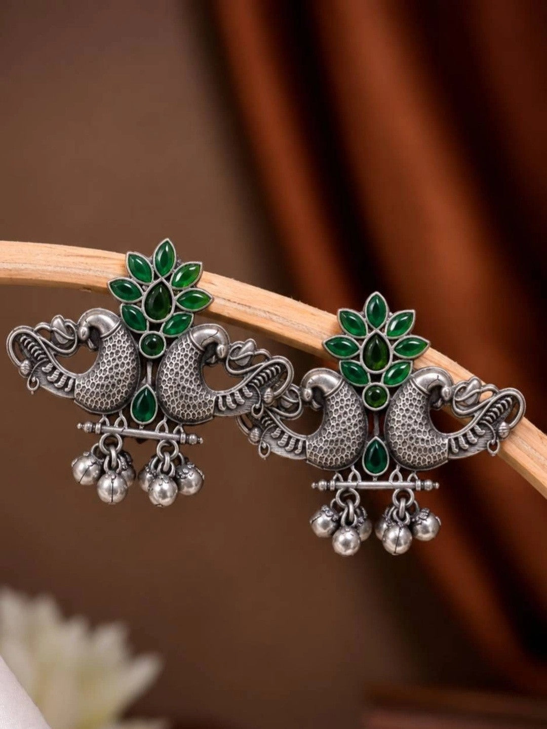 Pair of oxidised silver peacock stud earrings with green stones and silver ghungroo bells, displayed on a wooden prop against a brown draped background