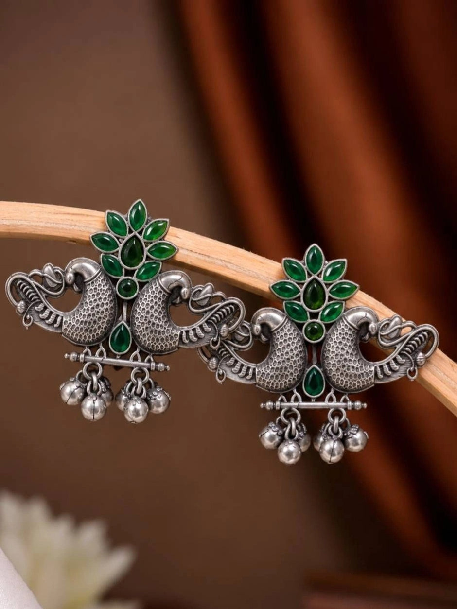 Pair of oxidised silver peacock stud earrings with green stones and silver ghungroo bells, displayed on a wooden prop against a brown draped background