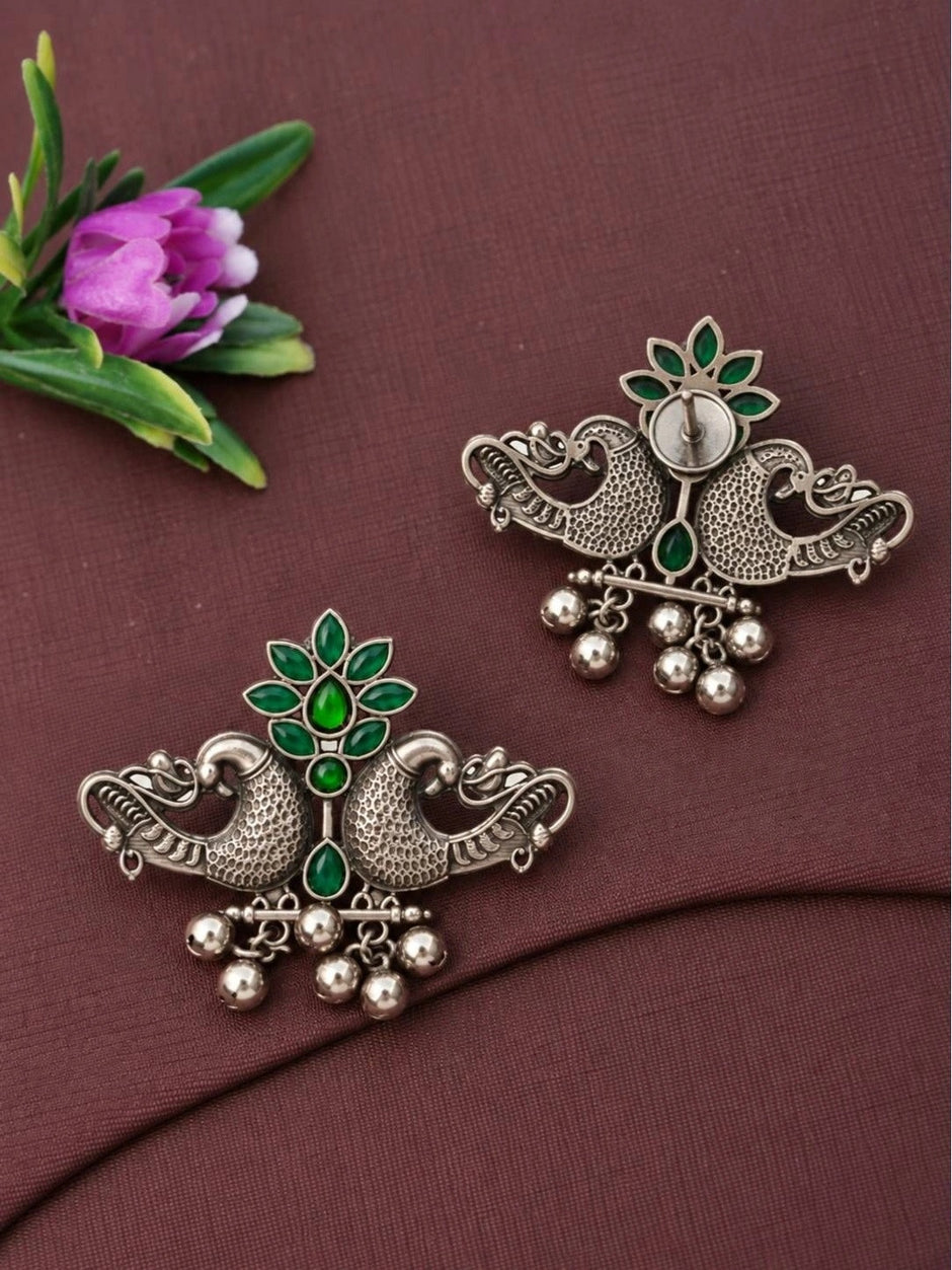 Front and back view of oxidised silver peacock earrings with green marquise stones laid flat on a mauve fabric surface beside a pink flower