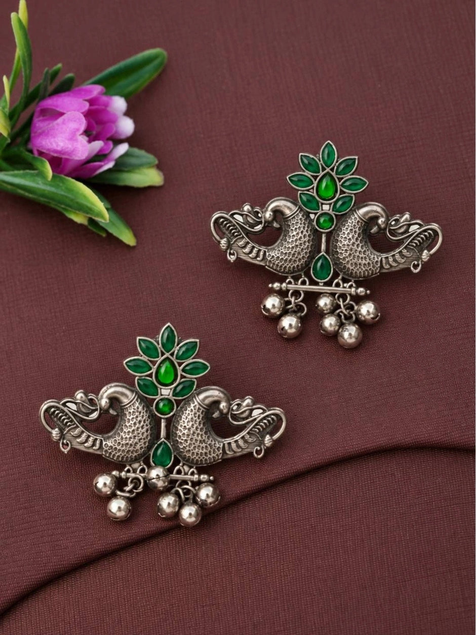 Pair of oxidised silver peacock stud earrings with green glass stones placed on mauve fabric next to a pink flower, showing full design detail