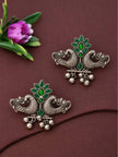 Pair of oxidised silver peacock stud earrings with green glass stones placed on mauve fabric next to a pink flower, showing full design detail