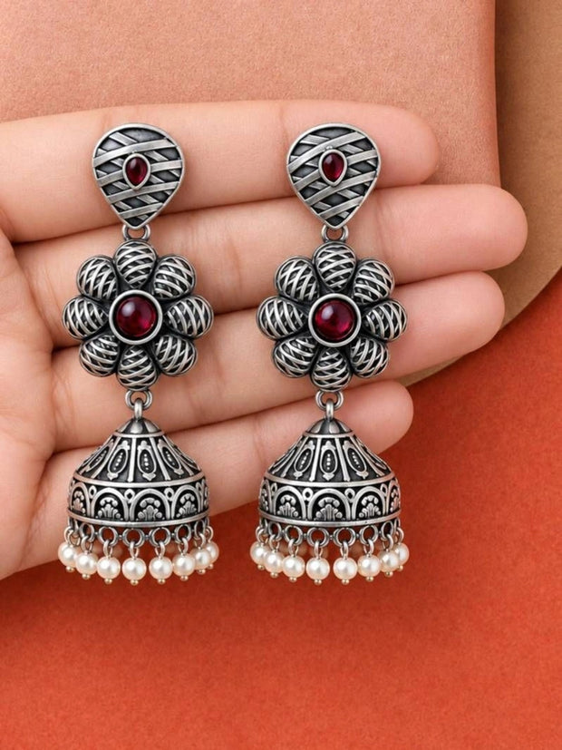 Pair of oxidised silver floral jhumka earrings with red stone and pearl fringe held in a palm
