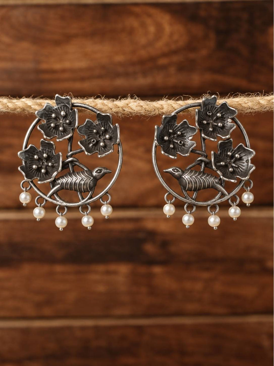 Pair of oxidised silver chandbali earrings with bird and floral motifs hanging from a rope against a wooden background