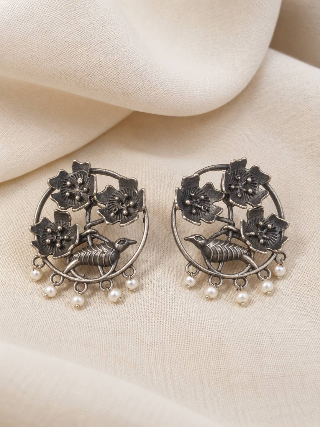 Front view of oxidised silver floral bird earrings with pearl drops laid flat on cream fabric