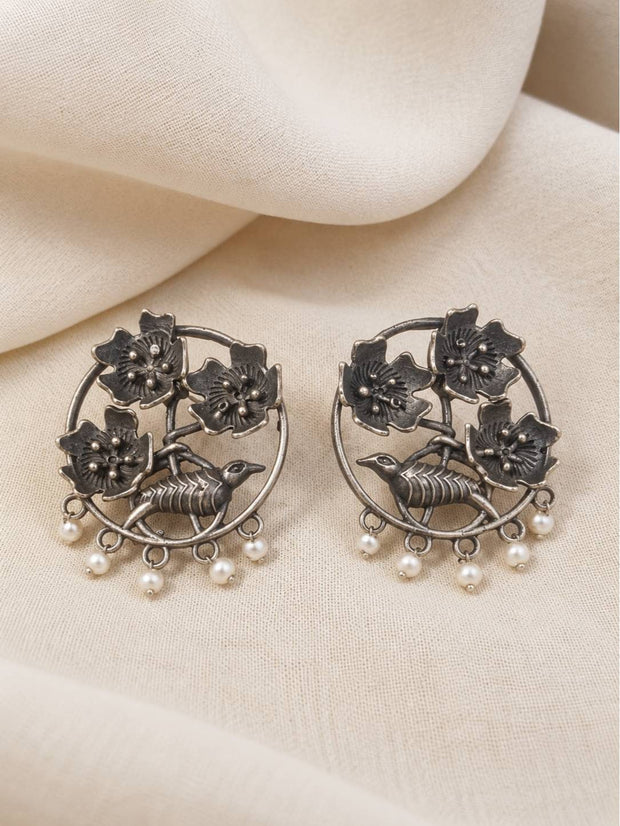 Front view of oxidised silver floral bird earrings with pearl drops laid flat on cream fabric