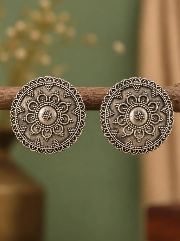 Pair of round oxidised silver mandala stud earrings hanging on a wooden branch against a green background