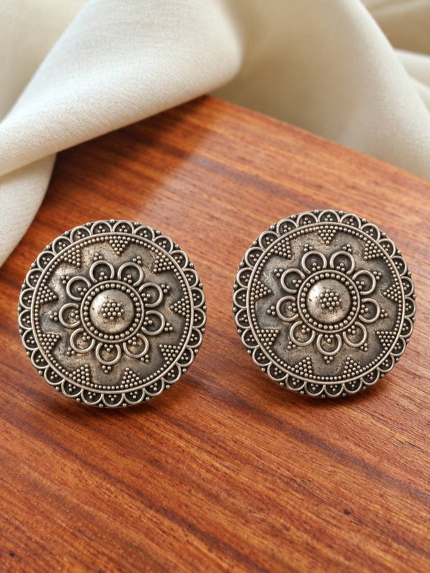 Two oxidised silver mandala stud earrings laid flat on a warm wooden surface showing intricate floral detailing