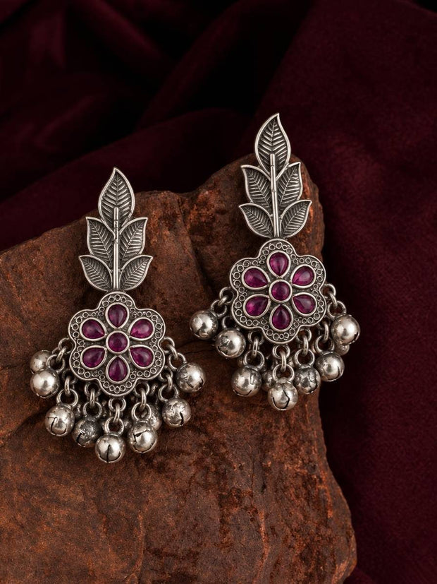 Pair of oxidised silver chandbali earrings with ruby red stones and ghungroo drops displayed on a rust-coloured rock against a dark red fabric background