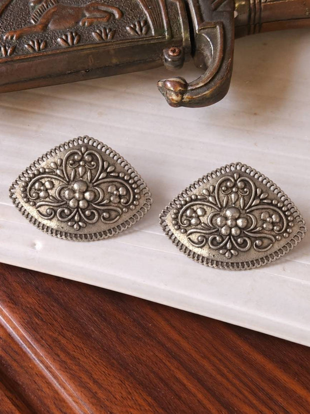 Two oxidised silver diamond-shaped stud earrings with embossed floral motif placed on white marble surface