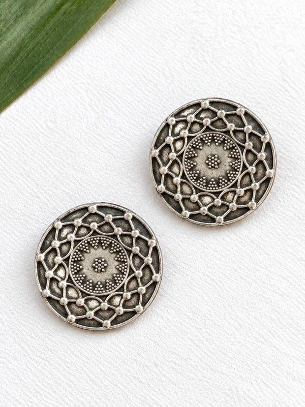 Two oxidised silver circular mandala stud earrings laid flat on a white surface beside a green leaf, showing front detail