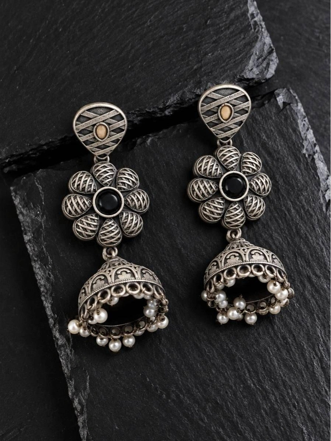 Two oxidised silver jhumka earrings with woven petal flower and pearl drop detail on slate