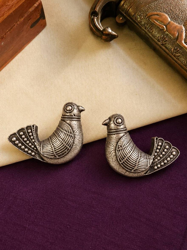 Two oxidised silver bird studs placed side by side on a cream and purple fabric surface with a decorative brass box in the background