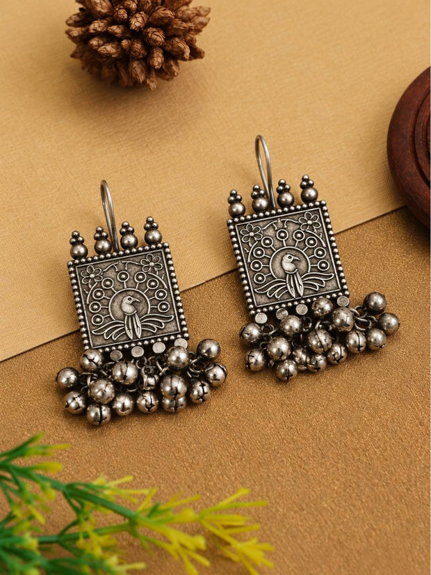 Front view of oxidised silver peacock earrings with floral engravings and ghungroo bells on a golden surface