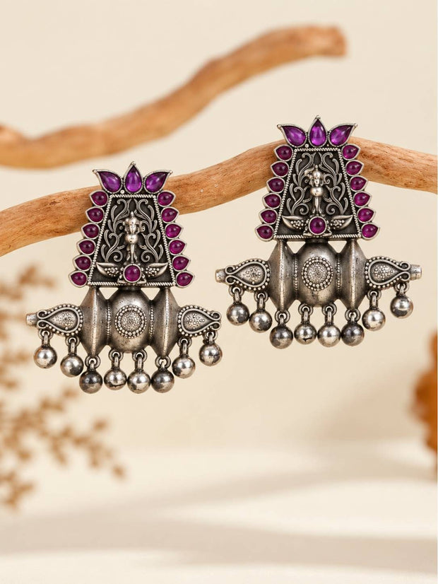 Pair of oxidised silver temple chandbali earrings with magenta stones and silver ghungroo ball drops, displayed hanging on a wooden branch
