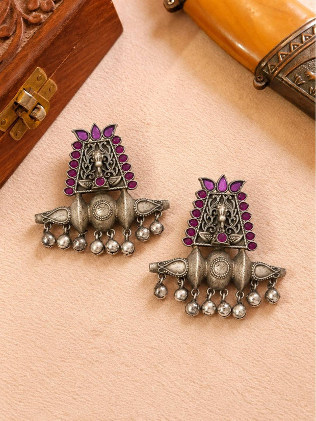 Front view of both oxidised silver temple earrings with deity motif and magenta stone border, laid flat on a cream surface near wooden boxes