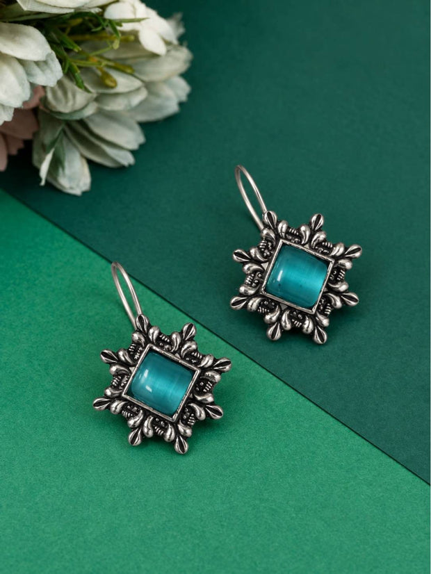 Pair of oxidised silver square drop earrings with teal cat-eye stone centres displayed on green surface