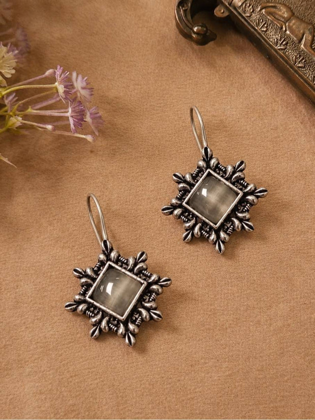 Two oxidised silver hook earrings with grey cabochon stones laid flat on a brown surface beside purple wildflowers