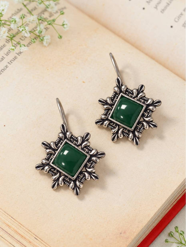 Two oxidised silver floral hook earrings with green stone centres laid flat on an open book with white flowers