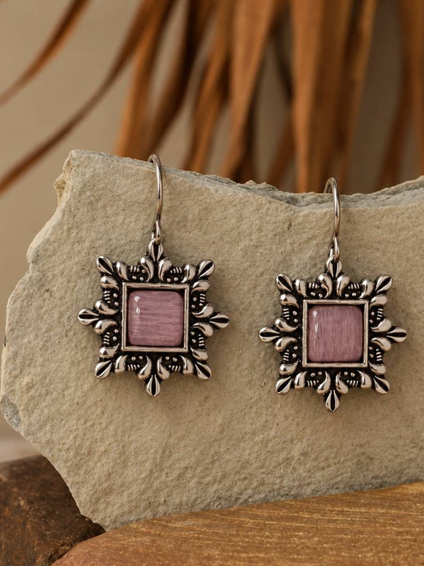 Pair of oxidised silver square drop earrings with pink cat-eye stone centres displayed on a rough stone surface