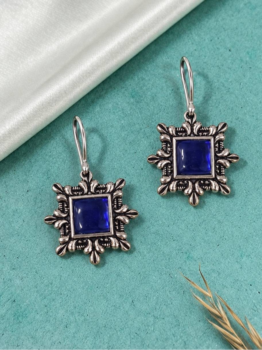 Two oxidised silver blue stone square earrings laid flat on a teal surface with dried grass