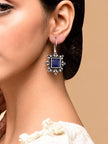 Woman wearing one oxidised silver square blue stone drop earring against a beige background