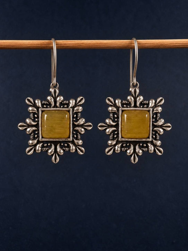 Pair of oxidised silver square drop earrings with yellow cabochon stone hung on a wooden rod against dark background