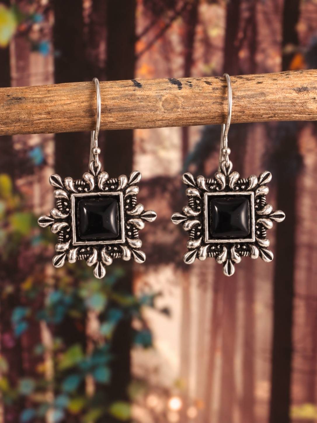Pair of oxidised silver square drop earrings with black stone centres hanging from a wooden stick