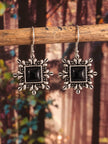 Pair of oxidised silver square drop earrings with black stone centres hanging from a wooden stick