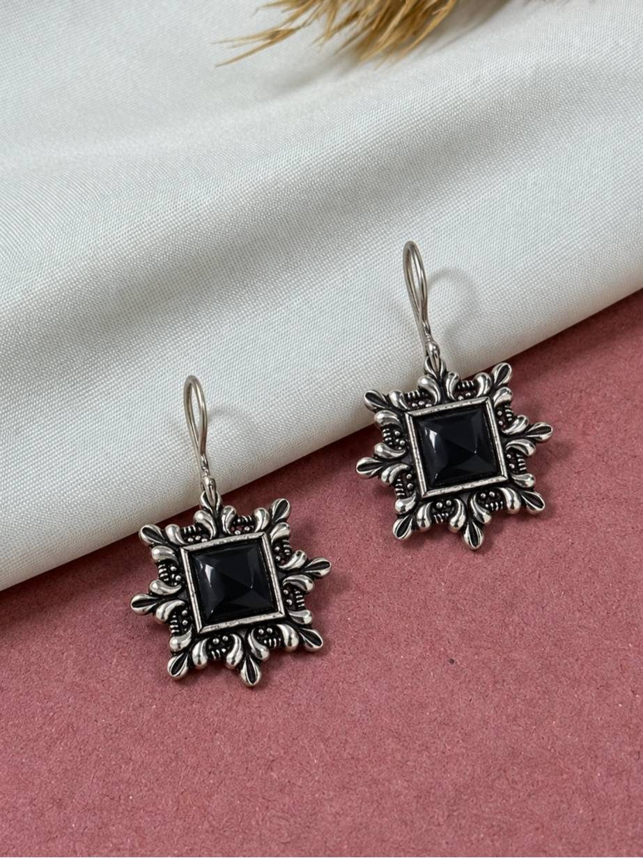 Two oxidised silver square earrings with floral border and black stone laid flat on a pink and white surface