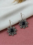 Two oxidised silver square earrings with floral border and black stone laid flat on a pink and white surface
