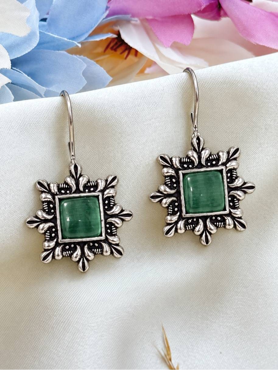 Pair of oxidised silver square drop earrings with green stone centre on white fabric with floral backdrop