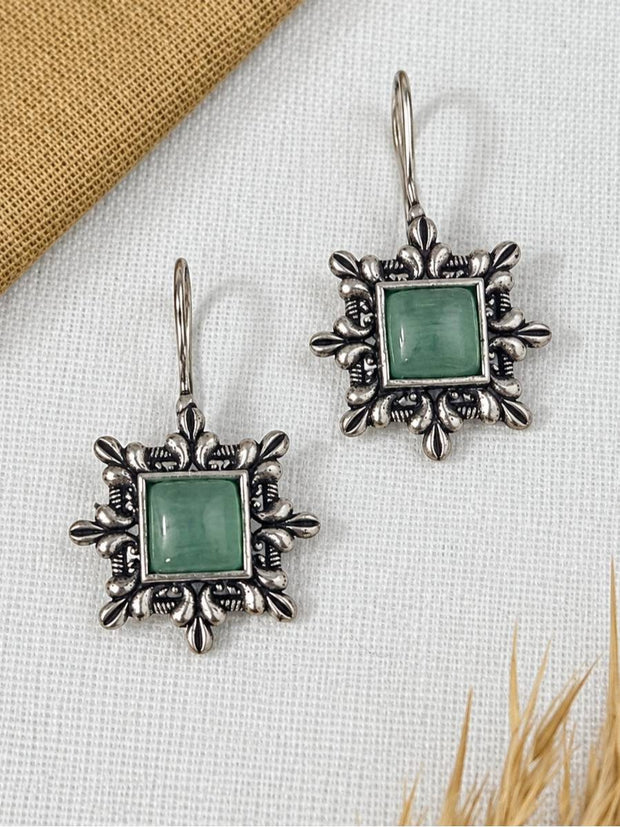 Two oxidised silver square earrings with ornate floral border and green cabochon stone on white linen