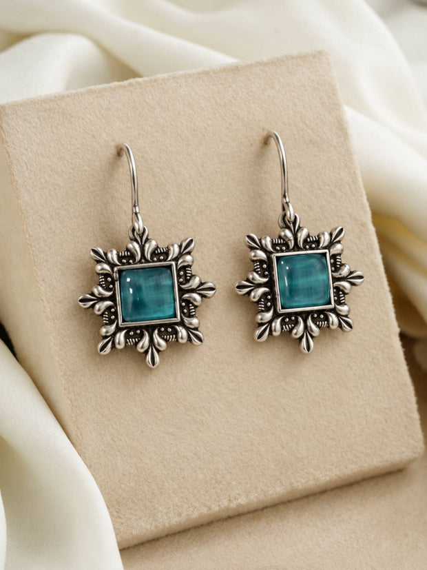Pair of oxidised silver square hook earrings with teal cabochon centre displayed on a beige jewellery box