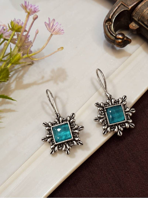 Front view of oxidised silver teal stone earrings laid flat on a white marble surface beside pink wildflowers