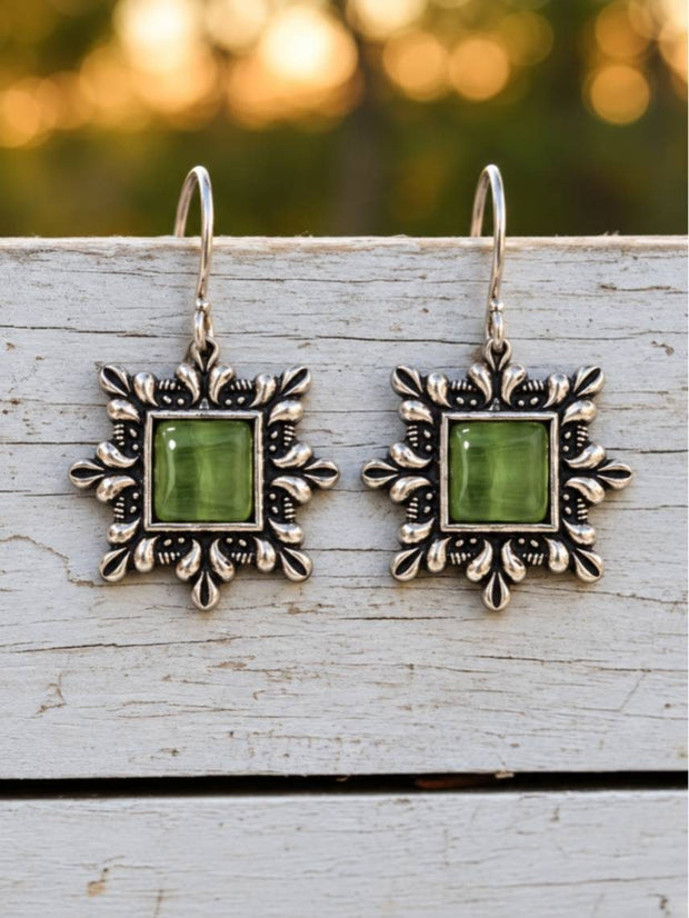 Pair of oxidised silver square drop earrings with green stone centres resting on a white wooden surface outdoors
