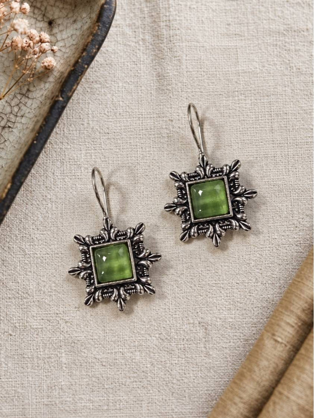 Two oxidised silver green stone drop earrings laid flat on a cream linen cloth beside a ceramic dish