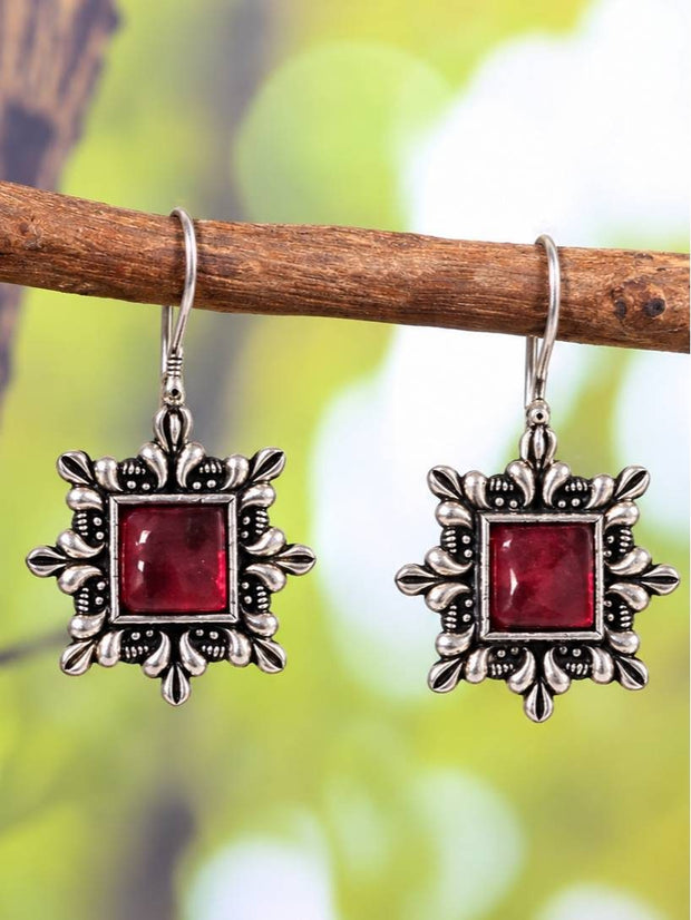 Pair of oxidised silver square drop earrings with ruby-red stone hanging on a wooden branch against a green background