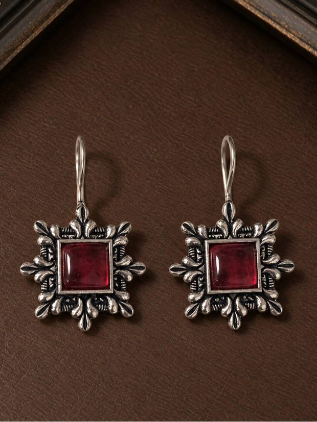 Front view of oxidised silver square drop earrings with red cabochon stone displayed on a dark brown leather surface