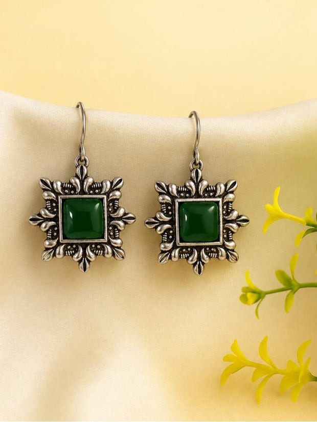 Pair of oxidised silver square earrings with green stone centres hanging on a cream display against yellow background