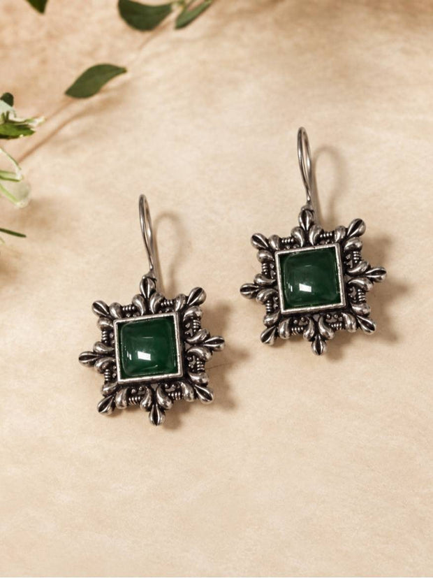 Two oxidised silver floral square hook earrings with green resin stones laid flat on a beige surface with green leaves